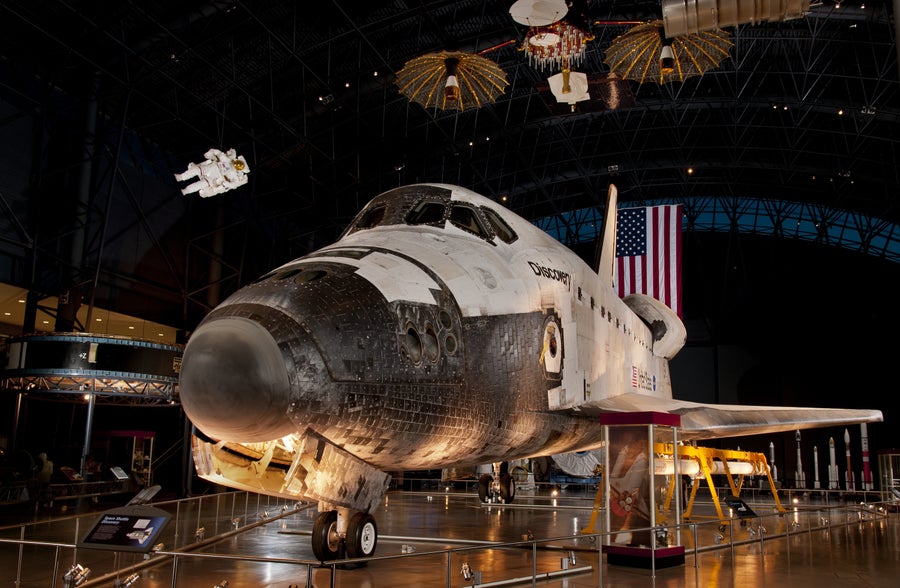 A Space Shuttle orbiter with a delta-winged spaceplane design, covered with white thermal protection blankets and black thermal protection tiles; marked Discovery on both sides of the forward fuselage.