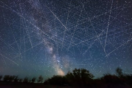 Long exposure photography composite showing light trails from low-Earth orbit satellites filling the sky, a treeline is in silhouette at the bottom of the frame