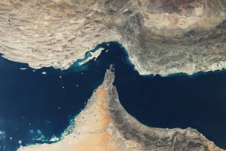 Satellite view of the Strait of Hormuz showing the narrow waterway between Iran (top) and the Musandam Peninsula of Oman and the UAE (bottom), with rugged desert mountains, sandy coastline, and deep blue waters dotted with small islands and reefs.