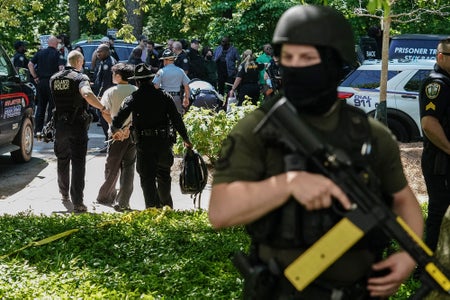 Police officers arrest a protester at Emory University in Atlanta, Georgia. In the right-hand foreground of the photo, out of focus, a police officer stands holding a semi-automatic pepperball rifle while wearing a black balaclava, leaving only his eyes visible