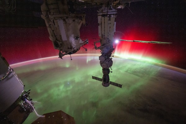 A green and red glowing aurora crowns Earth, with pieces of the International Space Station in the foreground