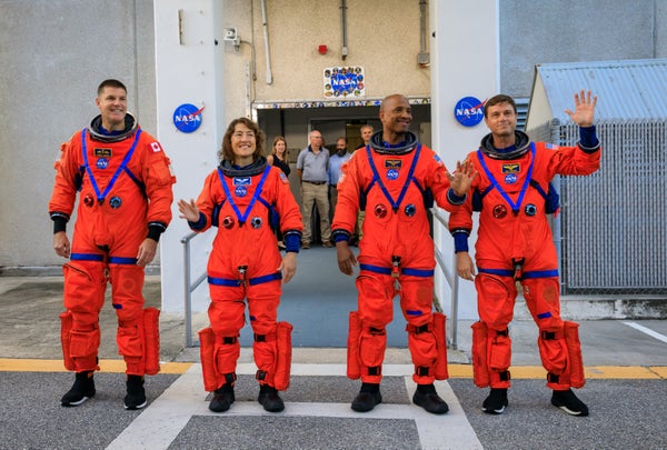 Four astronauts wearing orange spacesuits walk out of the Kennedy Space Center