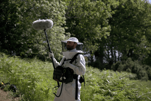 Un hombre vestido con un traje de apicultura se encuentra con un micrófono en un campo.