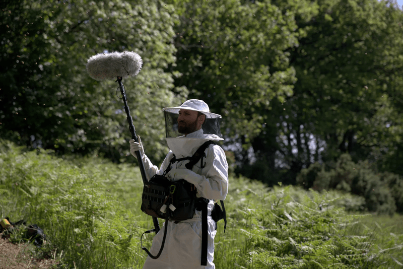 رجل يرتدي زي تربية النحل يقف حاملاً ميكروفونًا في أحد الحقول