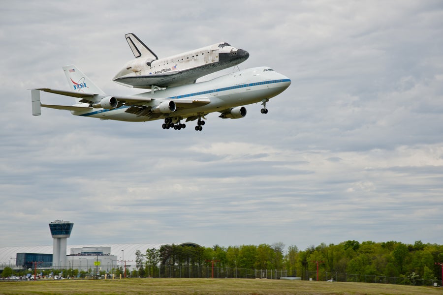 The space shuttle Discovery carried atop a Boeing 747 aircraft with an airfield tower in the background..