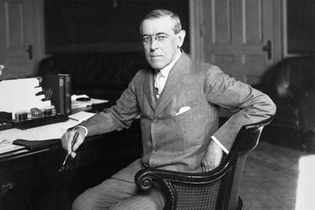 Black and white photograph of Woodrow Wilson sitting at a desk with a pen in his hand