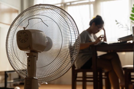 Unhappy Asian woman sitting in front of working fan suffering from heat in modern house on sunny summer day
