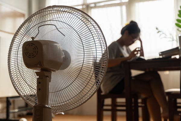 Unhappy Asian woman sitting in front of working fan suffering from heat in modern house on sunny summer day