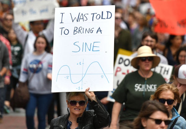 Woman at a protest march, holding a sign that says "I was told to bring a sine" with a sine wave pitcured