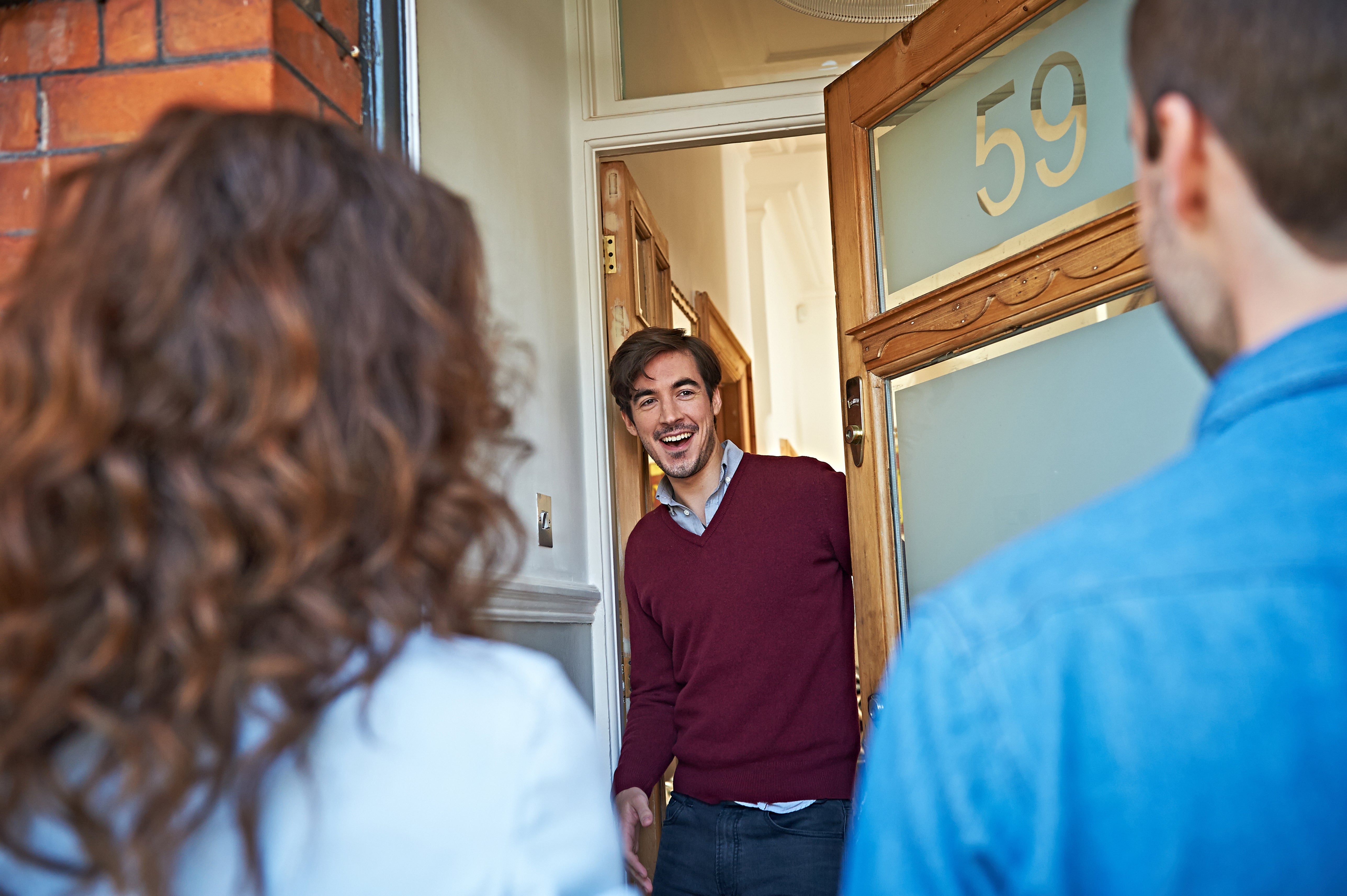 Young man with red sweater at his front door being surprised by two friends (backview).