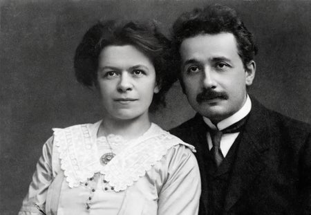 B/w portrait of Albert Einstein in black suit and tie and his wife Mileva Marić Einstein wearing a white top with lace collar and necklace with round pendant.