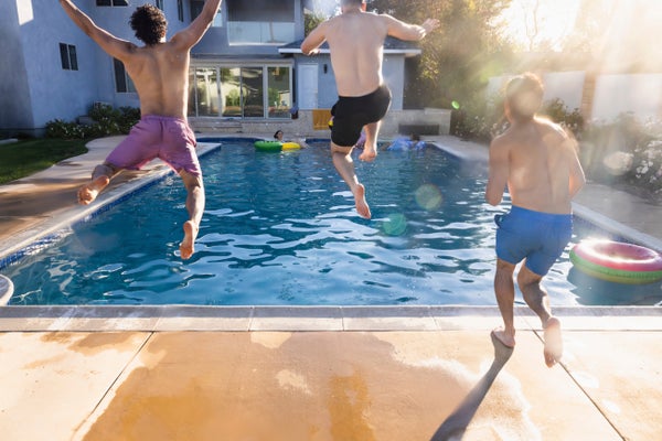 Rear view of 3 male friends in purple, black and blue swim trunks jumping into swimming pool together