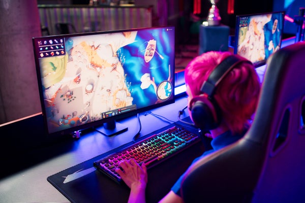 Rear view of a gamer with pink hair wearing headset and talking into the microphone to communicate while playing a game on a desktop with colorful ambient lighting and a rainbow backlit keyboard