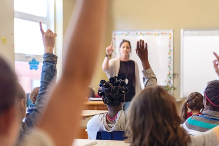 Grade school aged students raising their hands in front of a teacher in a school classroom