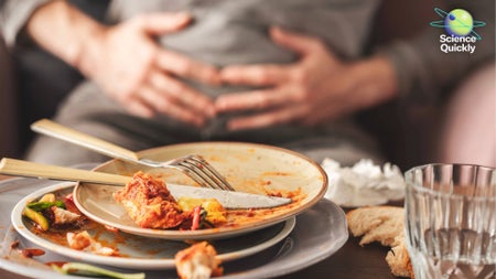 An image of a person holding their bloated stomach with a pile of plates in front of them on a table with food scraps left on them signifying the end of a large meal