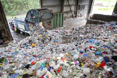 A recycling truck dumps recyclable items onto the floor of a recycling plant, which is covered with more plastic and other items