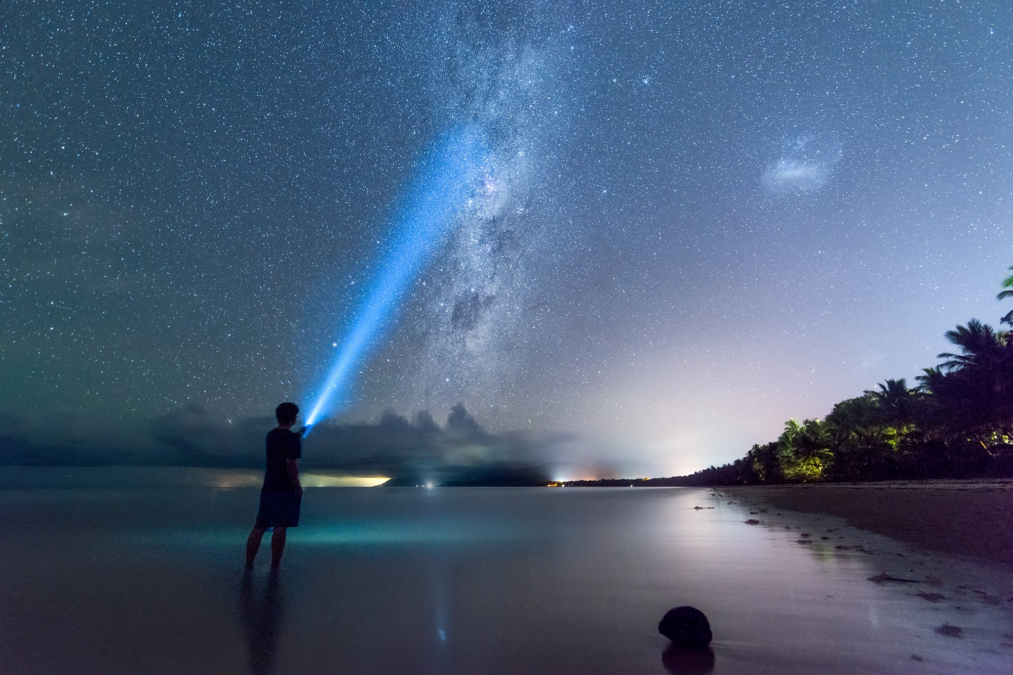 A person holding a flashlight stands on a beach while viewing the Milky Way in the night sky