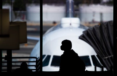 silhouette of a person wearing a face mask stands at an airport, with an airplane visible in the background.
