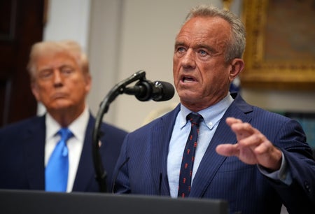Robert F. Kennedy, Jr. stands at a podium speaking with Donald Trump in the background.