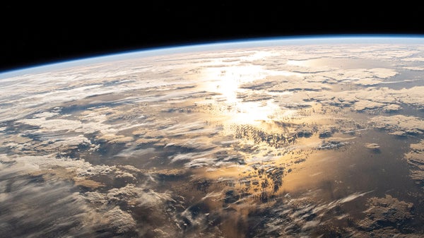 View of Earth from space, with a curved view of the horizon. In the foreground, sun reflects off the Atlantic Ocean between clouds.