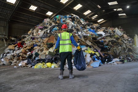 A man stands in a high-viz vest facing away from the camera in front of a pile of trash in a large hangar-like room