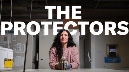 A white woman with long curly brown hair wears a pink shirt and large, thick gloves. She holds her hands folded on a table in front of her and stands in front of large tan freezers. She is looking just above the camera lens, and large text that says "The Protectors" sits above her head.