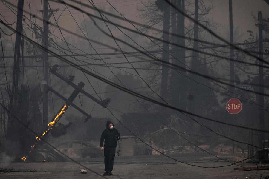 A masked person walks through Altadena's devastated business district through a crisscrossing web of downed power lines and haze of smoke on Wednesday, January 8, 2025 during the Eaton fire