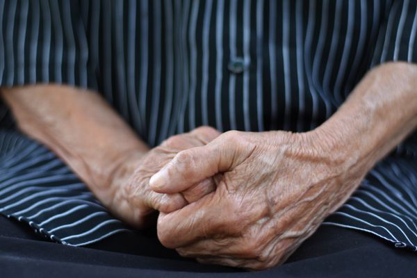 The midsection of an older woman who holds her hands in hand in her lap and wears black short with white straps.