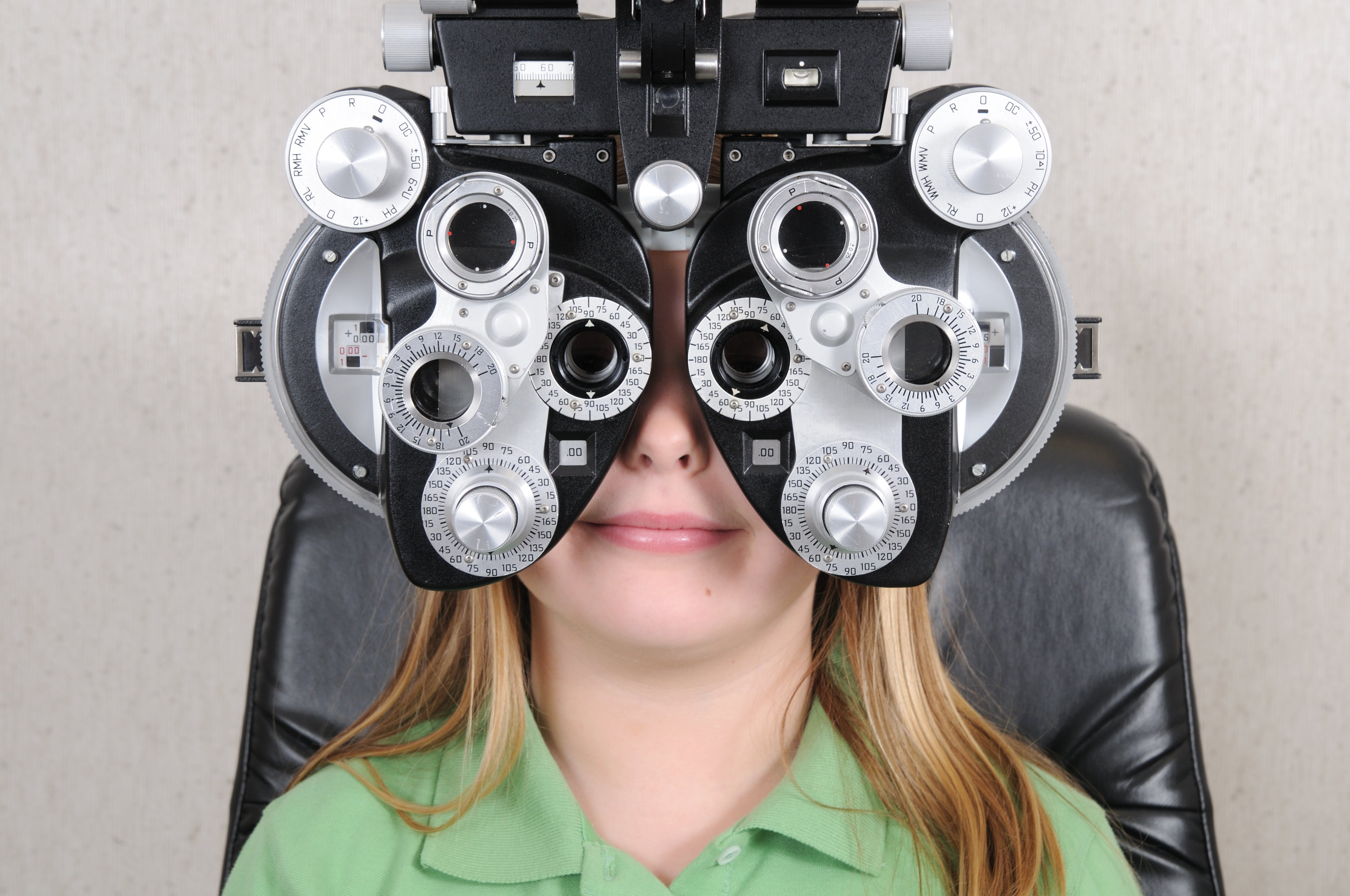 Young girl with blond hair and green short getting eye exam with phoropter.