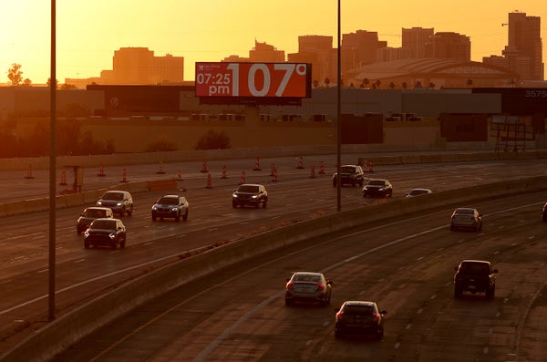 A billboard over a highway reads 107 degrees fahrenheit with an orange sky and part of the Phoenix skyline in the background