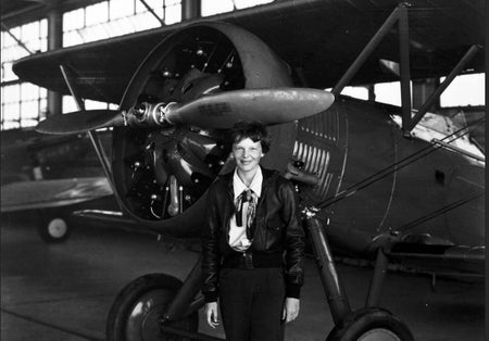 Amelia Earhart stands in front of a plane in a black-and-white photograph