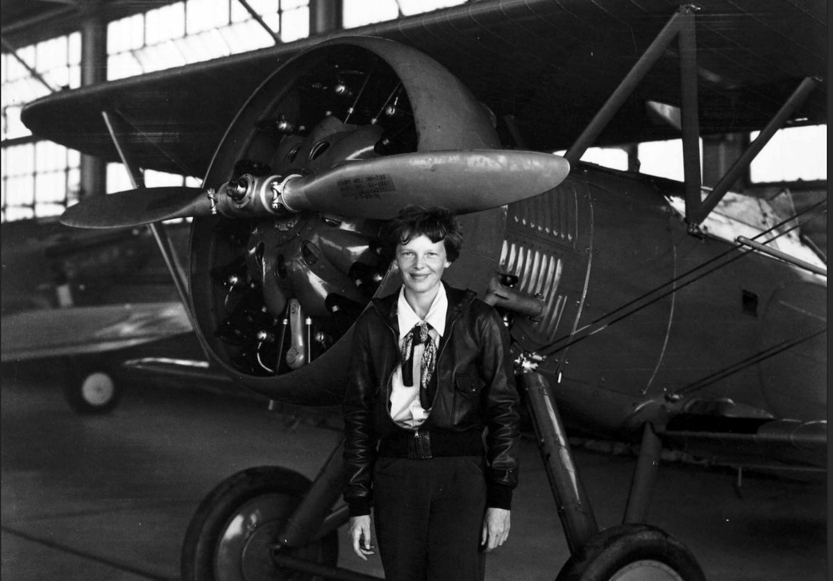 Amelia Earhart stands in front of a plane in a black-and-white photograph