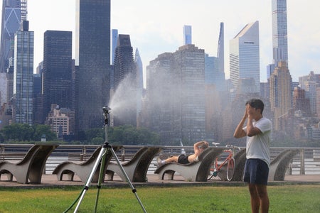 An adult stands with their hands covering their face in the stream of a large sprinkler set up on the grass in a park on the East River in Long Island City with the Manhattan skyline in the background
