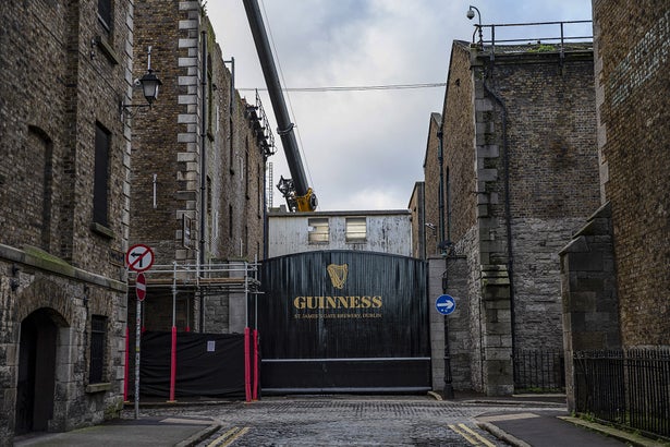 Photograph of a view looking down a narrow street in Dublin, Ireland towards a closed black gate between two brick factory buildings at the Guinness Saint James Gate Brewery. The gate has the Guinness logo in gold paint displayed on it.