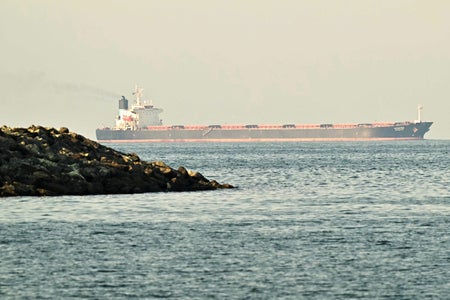 Large oil tanker traveling across calm water near a rocky breakwater, with a faint plume of exhaust rising from the ship’s stack under a hazy sky.