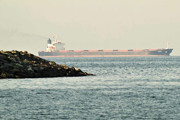 Large oil tanker traveling across calm water near a rocky breakwater, with a faint plume of exhaust rising from the ship’s stack under a hazy sky.