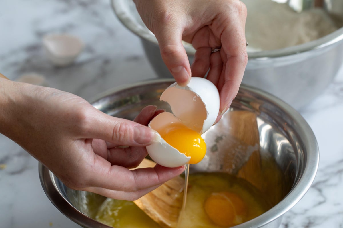 Person's hands cracking an egg open with the yellow yolk over a round metal mixing bowl with natural light