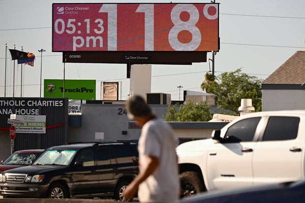 Photograph of a tall LED billboard reading the time of 5:13 PM and temperature of 118 degrees Fahrenheit. In the foreground a man is looking towards the billboard while walking past cars in a parking lot