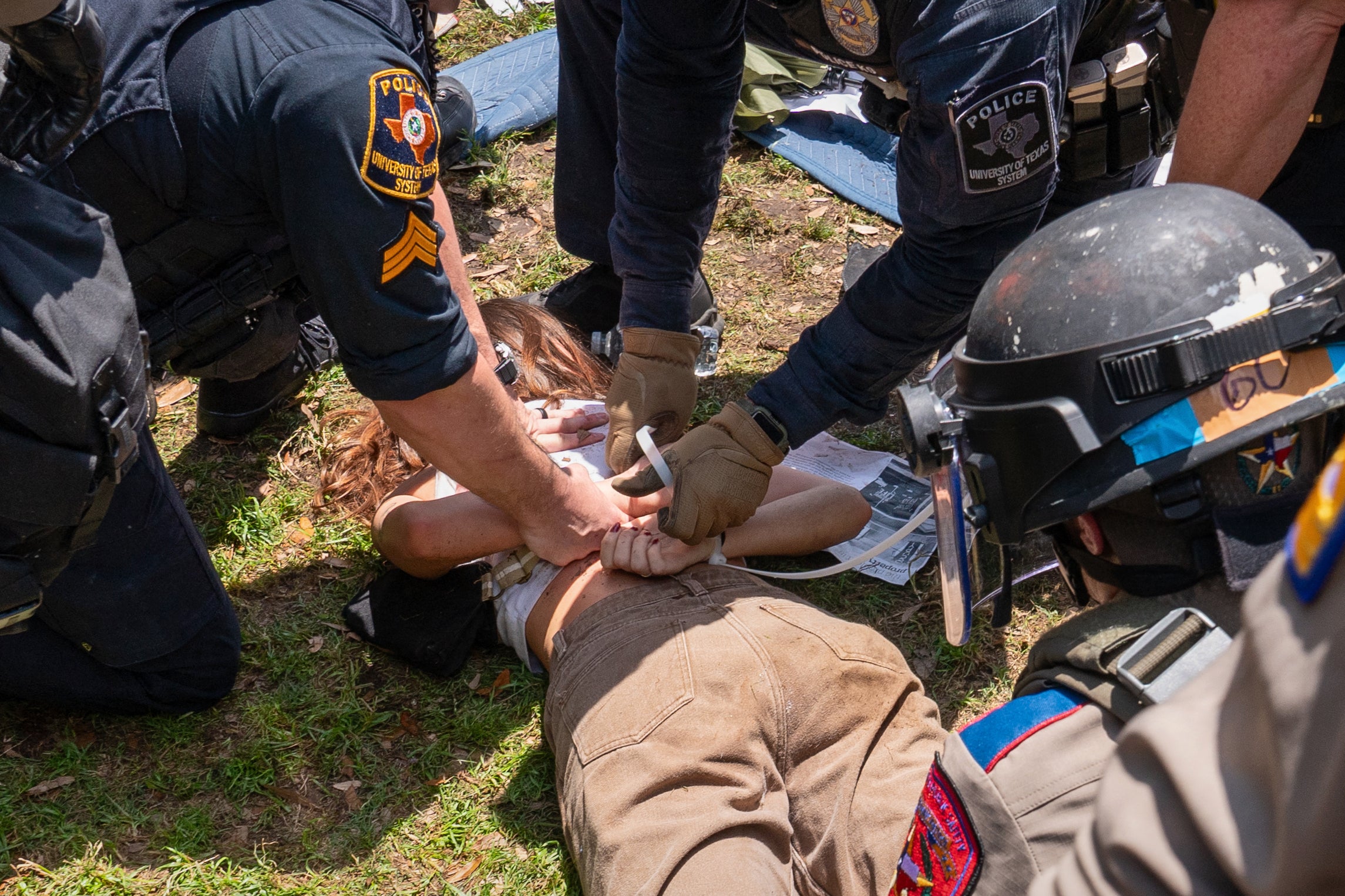 Three (3) law enforcement officers hold a protestor on the ground in a face down position as the officers bind the student’s hands behind their back with zipties