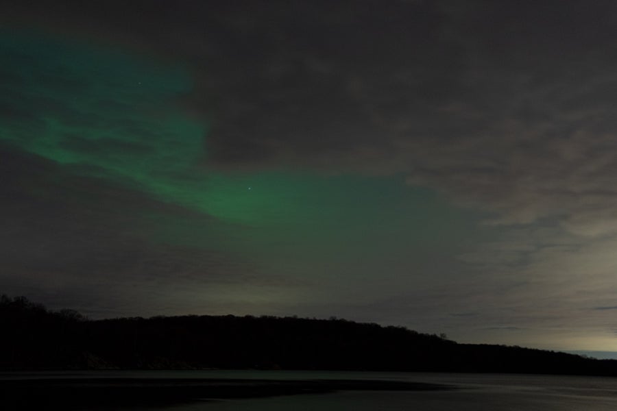 Faint pink and green auroras seen in a cloudy sky with the dark ground below.