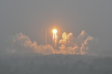 Rocket lifting off from launch pad with white smoke and light flare at center.