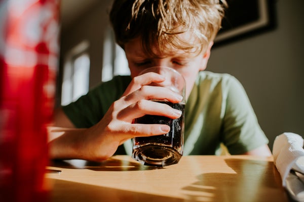 A child holds and sips from a glass full of fizzy cola in a sunny room