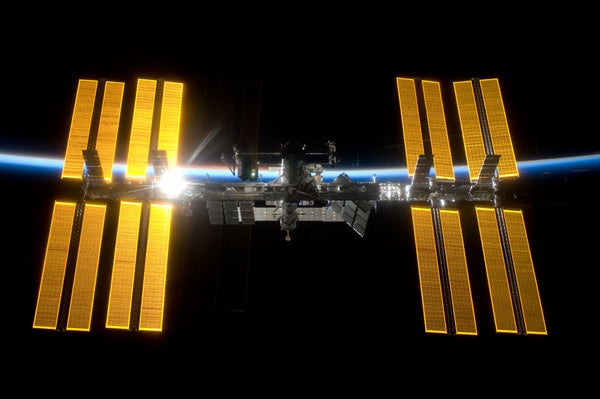 The International Space Station (ISS) backdropped by the blackness of space and the thin line of Earth's atmosphere, the sun shining through the solar array to the left of center