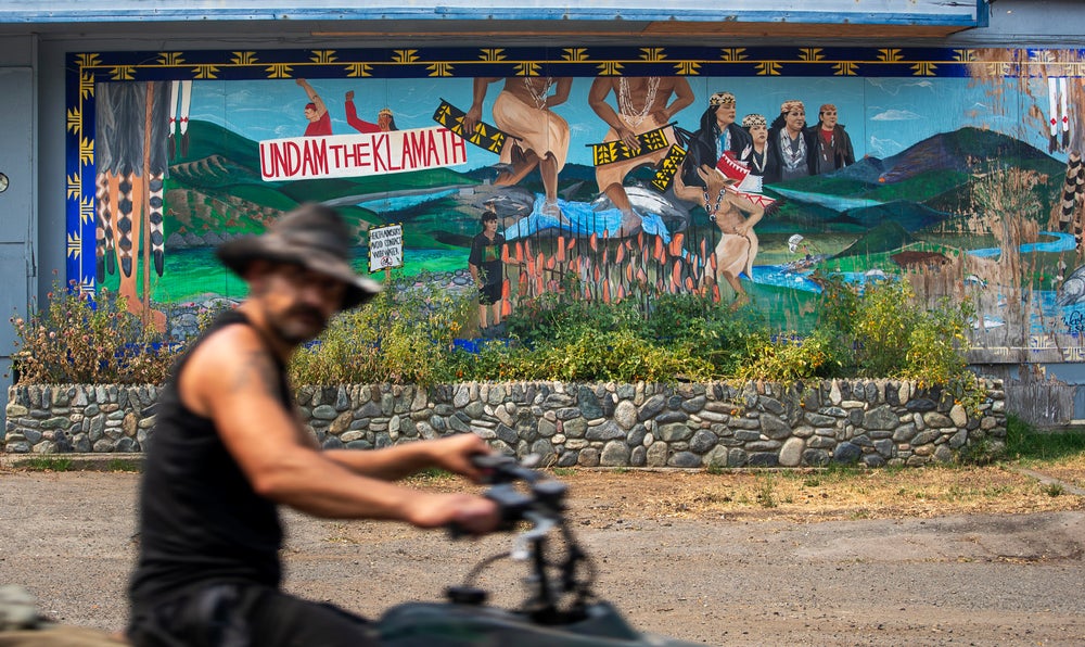 Un hombre pasa en motocicleta junto a un mural que dice:
