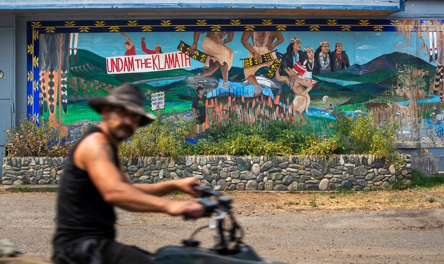 A man rides a motor bike past a mural that reads, "Undam the Klamath," advocating for the removal of the dams on the Klamath River with imagery depicting the past industry along the river, the restoration, and the future history of the river after removal. In the center Northern California Native people people perform a stomp dance to bring balance to the world