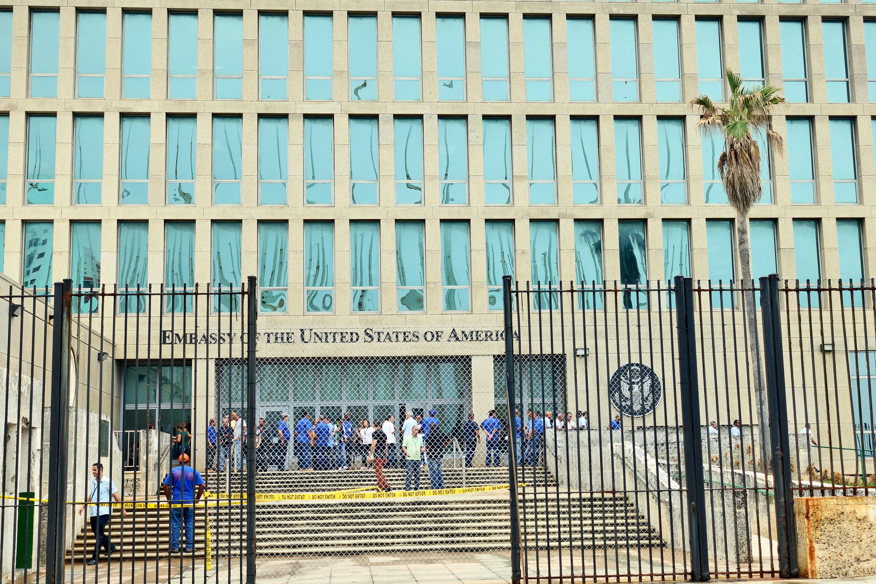 A view of personnel in front of the entrance to the U.S. Embassy in Havana, Cuba, looking through an open black metal bar security gate, chain link fence, and caution tape