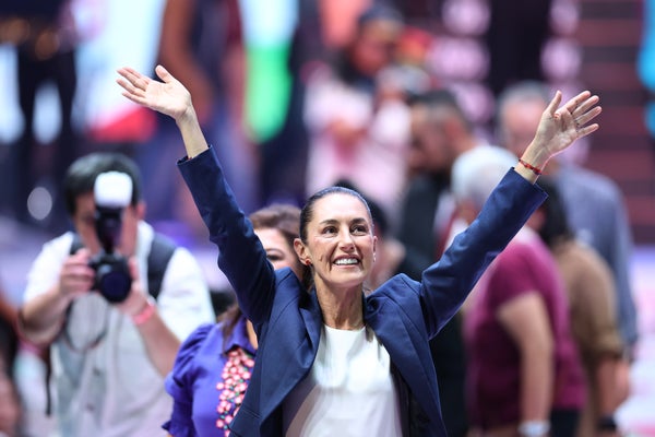 President-elect Claudia Sheinbaum in blue jacket hands up in celebration and crowd and photographer in background.