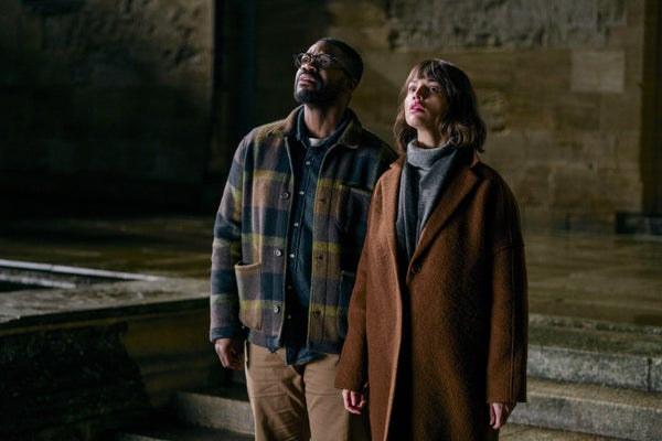 Male in plaid shirty and female in brown coat standing in empty space looking up.