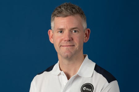 Portrait photograph of John McFall, member of the ESA Astronaut Class of 2022, in front of a blue backdrop