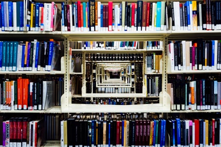 A library shelf packed with books, with a rectangular hole cut out of the center, revealing infinitely more shelfs behind.